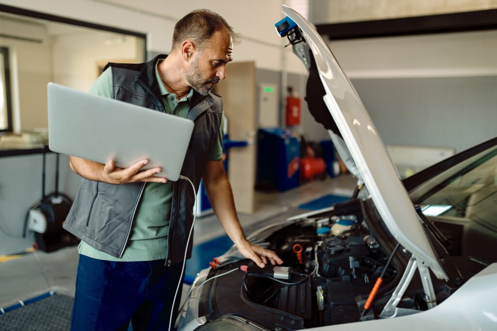Auto repairman using laptop while examining car engine in a work