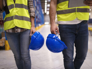 Two unrecognizable workers in reflective suit walking through warehouse and holding blue protective hardhats.