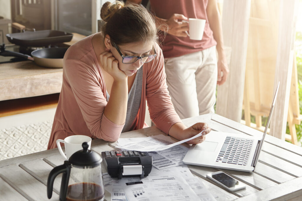 Young housewife dressed casually focused on paperwork, reading document in her hands with concentrated look, managing domestic finances alone while her husband drinking coffee in background