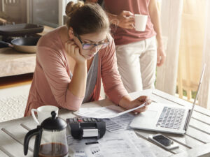 Young housewife dressed casually focused on paperwork, reading document in her hands with concentrated look, managing domestic finances alone while her husband drinking coffee in background